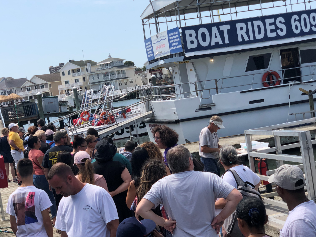 People line up for boat rides on a sunny day at a pier with a sign that reads 