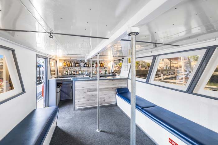 Interior of a boat with a bar. Blue seating lines the walls. A bar with bottles and glasses is visible.