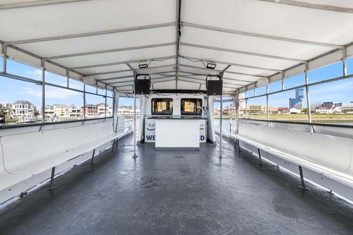 Interior of a boat with white benches, bar, and open windows offering a view of buildings and sky.