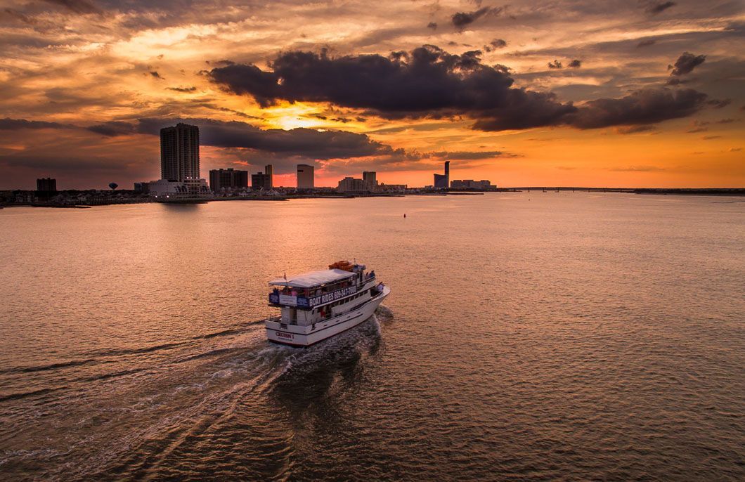 Boat sails across water toward a city skyline at sunset. Orange and gold sky.