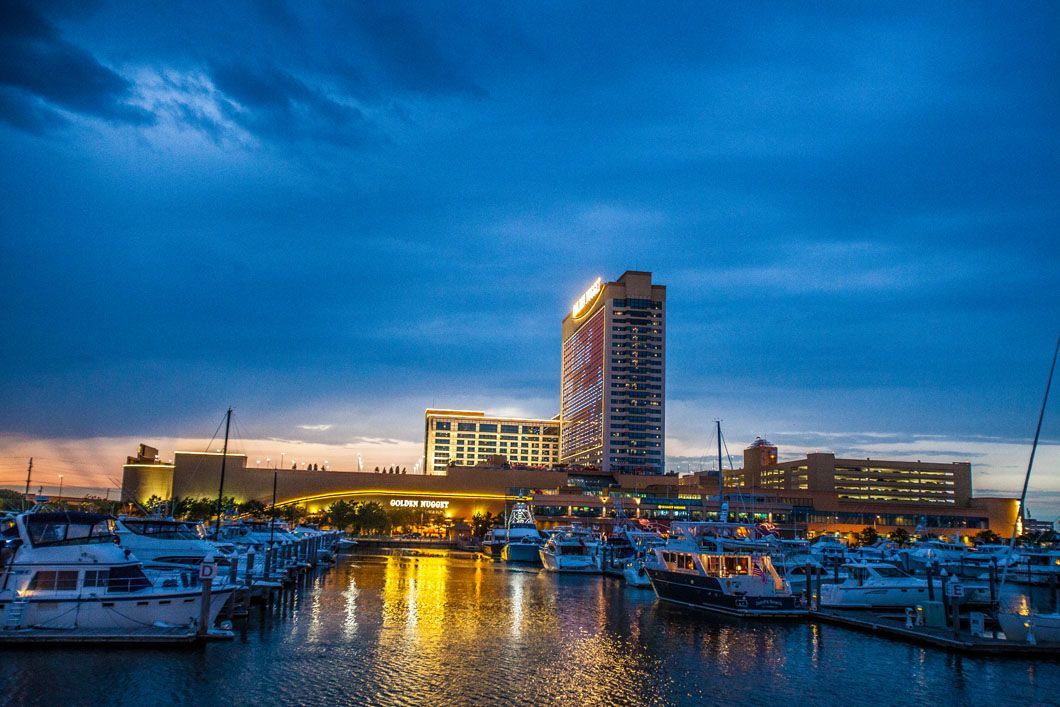 Marina at dusk with boats, a lit waterfront, and a tall building under a dark blue sky.