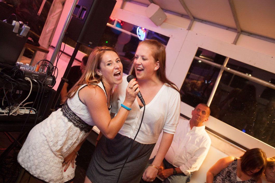 Two women singing karaoke, one holding the microphone, in a brightly lit room.