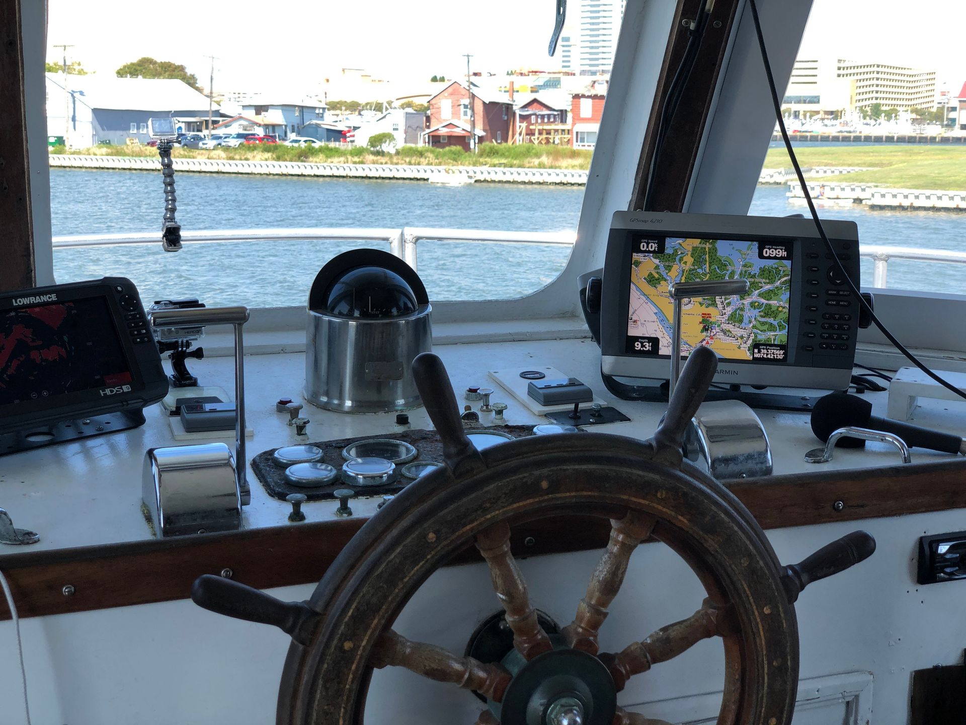 View from the helm of a boat, showing the steering wheel, navigation display, and buildings on the shore.