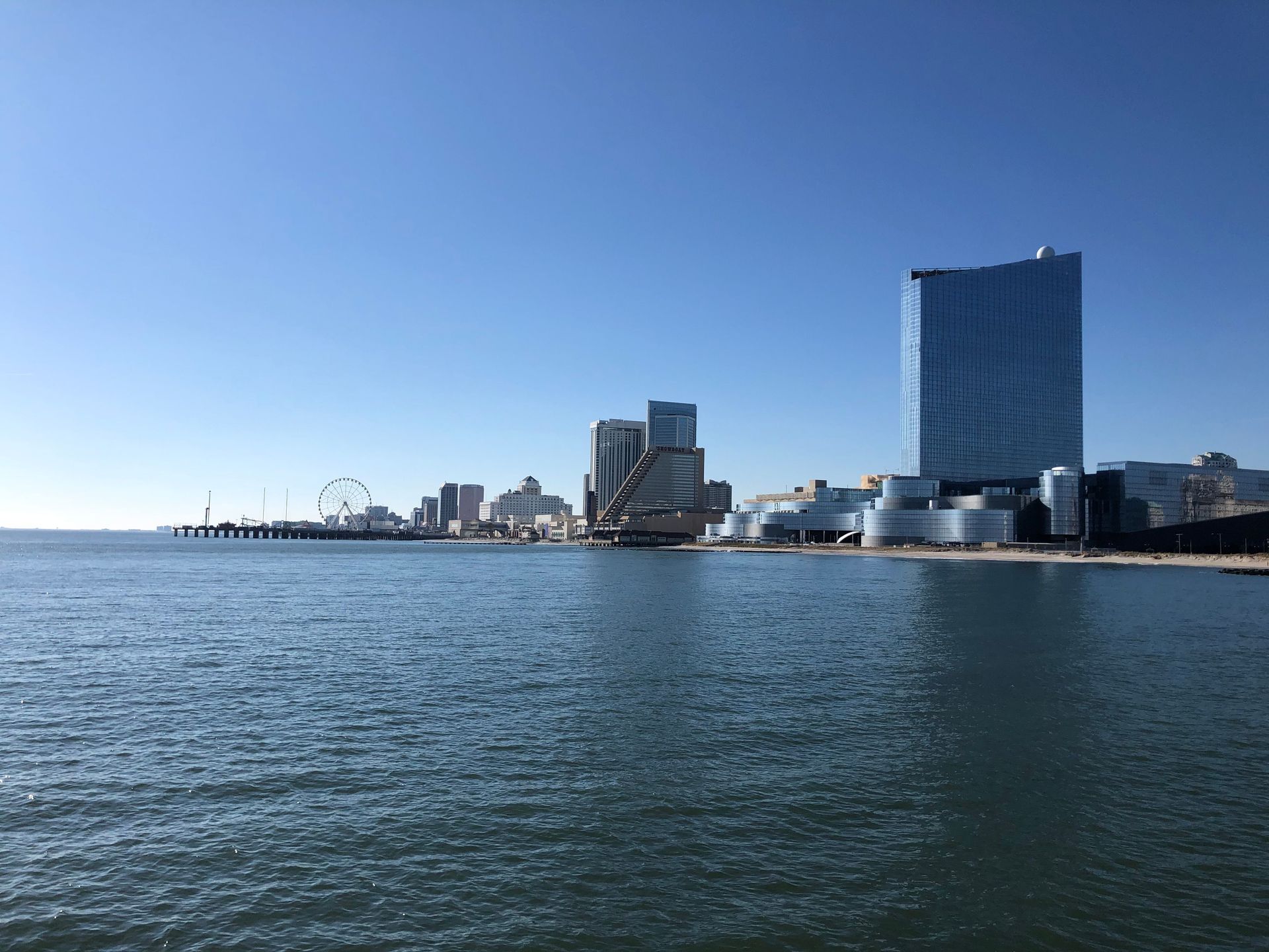 City skyline along a blue ocean under a clear sky. Several buildings, including a tall blue tower, are visible.