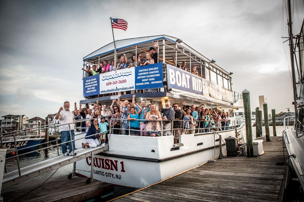A two-story boat, "Cruisn 1," loaded with people waving, docked at a pier, under a cloudy sky.