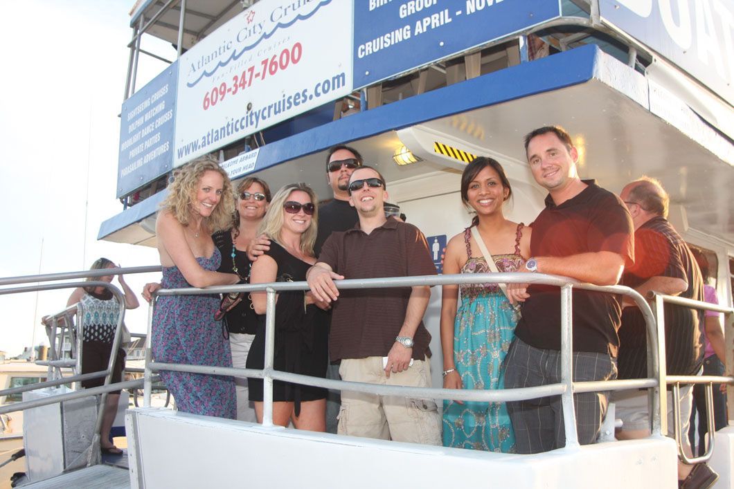 Group of people on a boat deck smiling near a railing. Blue and white structure, sunny background.