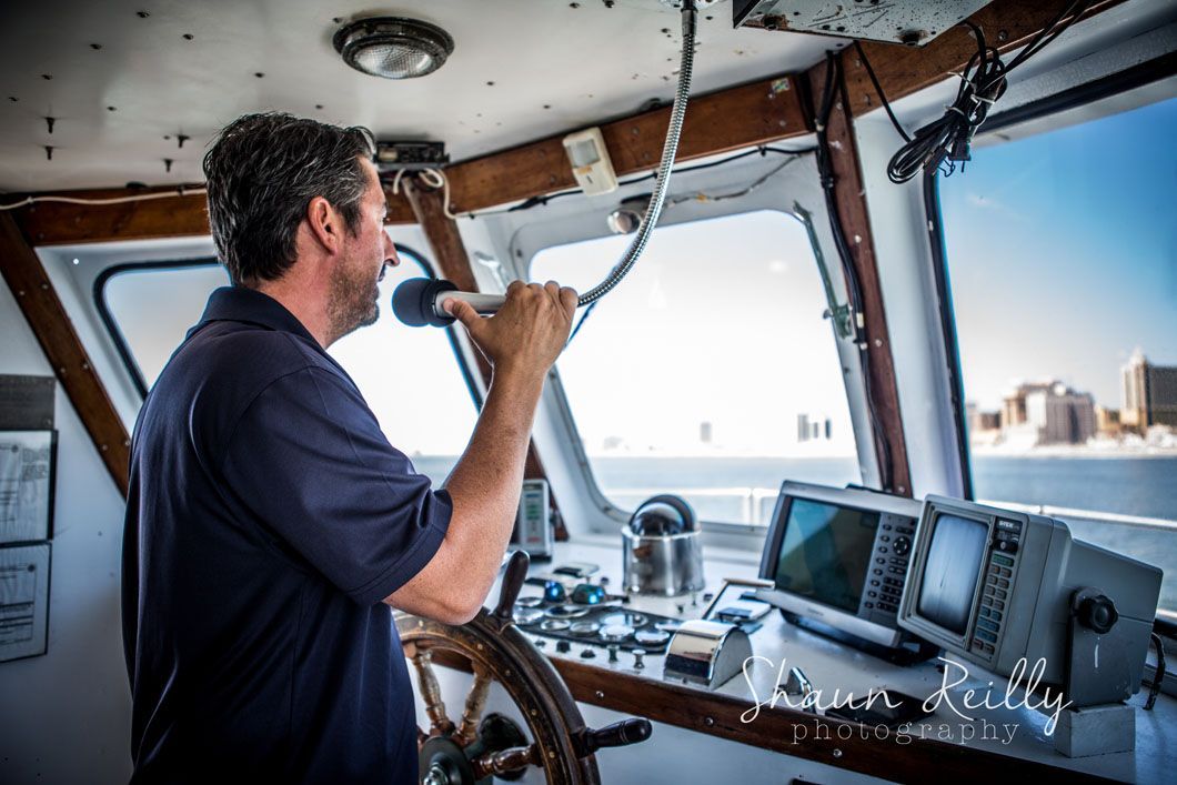 A man steering a boat, holding the wheel, inside the boat's cabin, with cityscape in the distance.