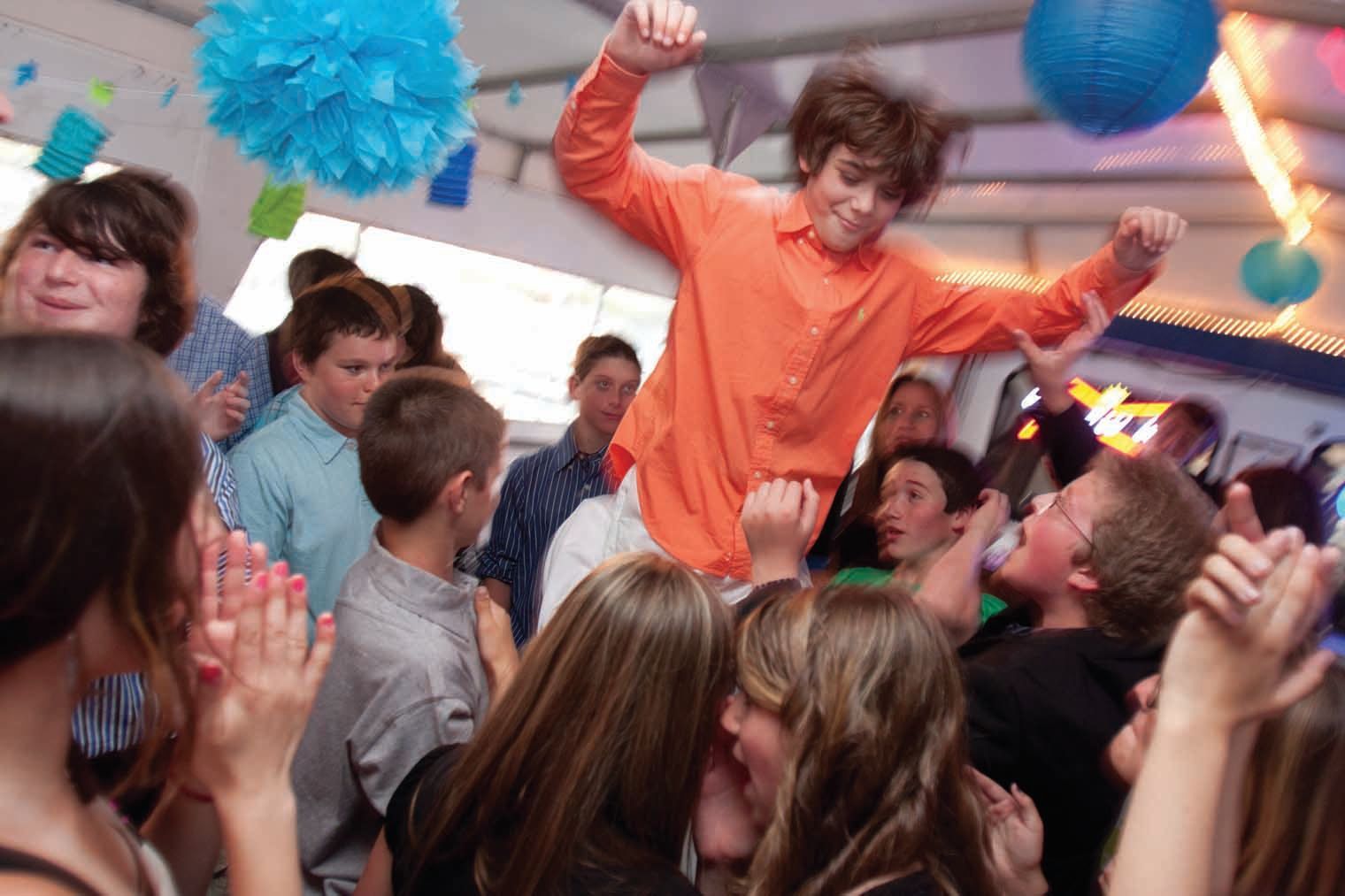 Teen crowd at a party, lifting a person with an orange shirt; confetti and decorations.