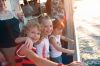 Three children smile from a boat railing, ocean in the background. Sunny day, bright light.