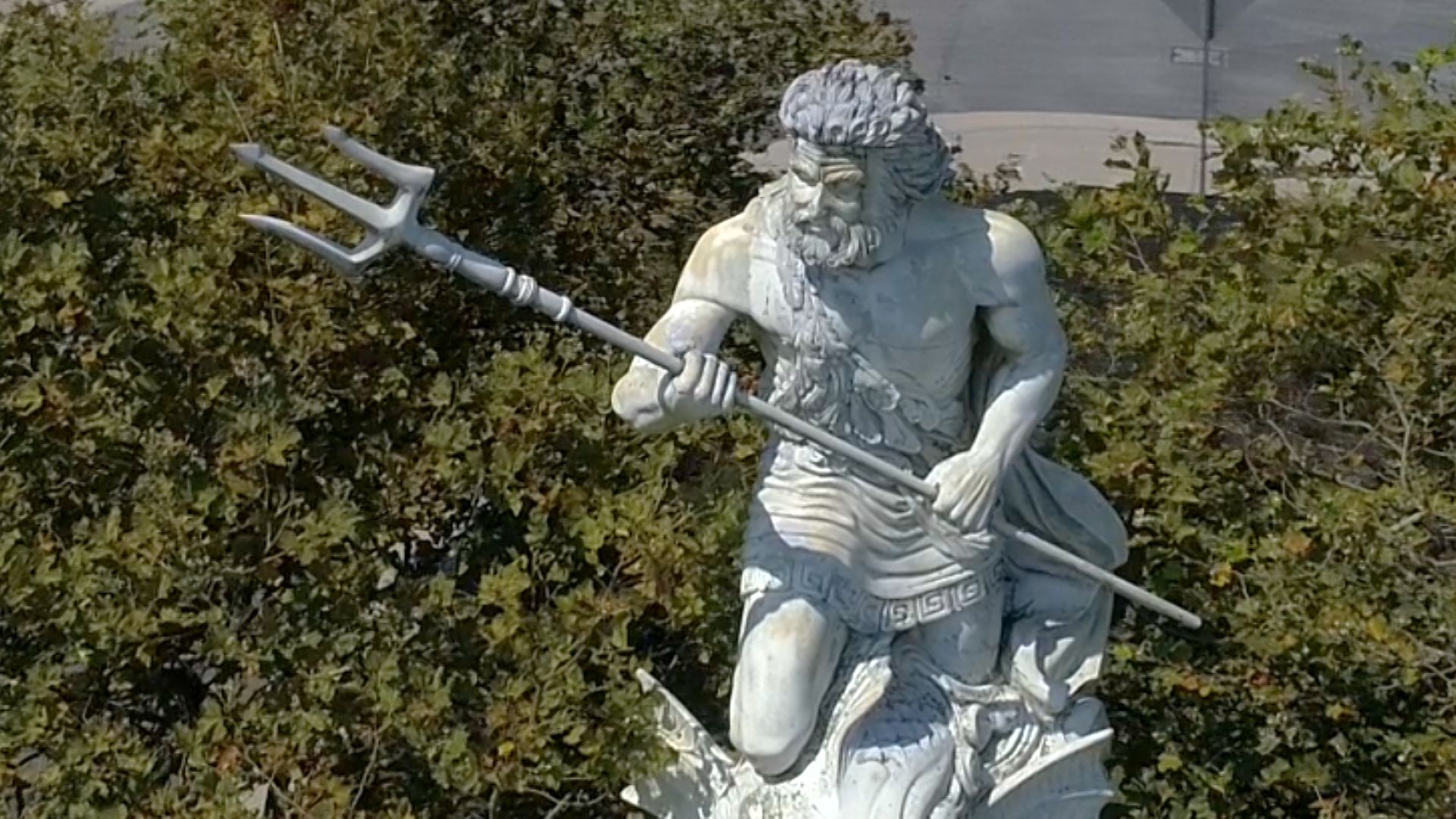 Statue of Neptune, a bearded man, holding a trident. The statue is white with a green backdrop.