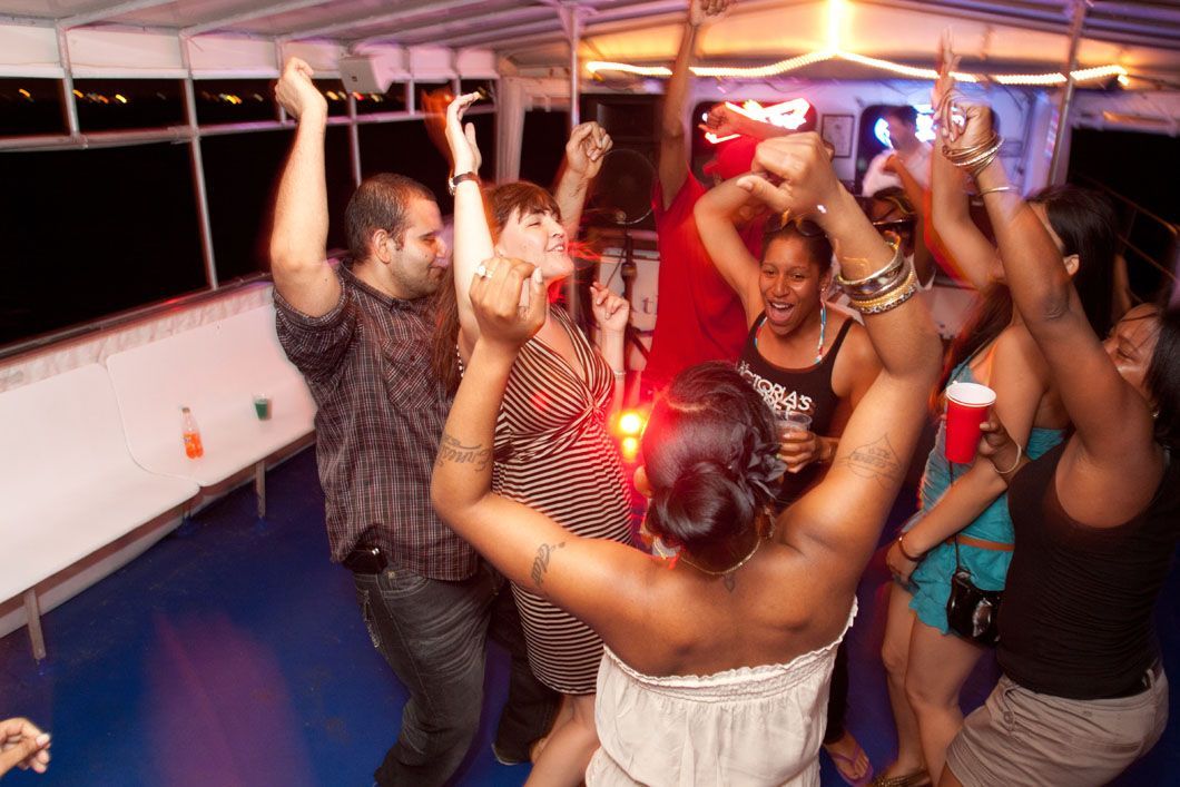 People dancing on a boat deck at night, raising arms, celebrating. Bright lighting.