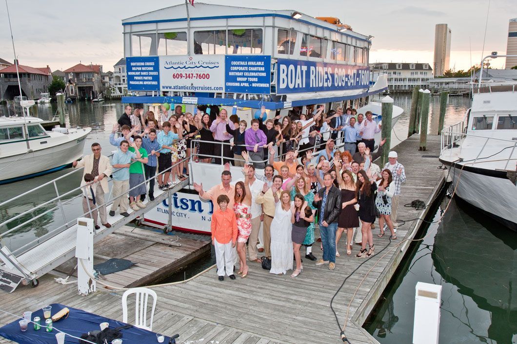 Large group of people on a dock next to a two-story boat. Building in background.