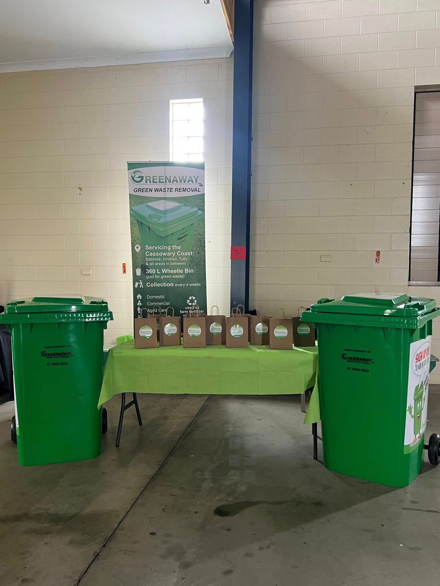 Two green trash bins flank a green table with brown paper bags and a banner, indoors.— Greenaway Green Waste Removal in O'Briens Hill, QLD