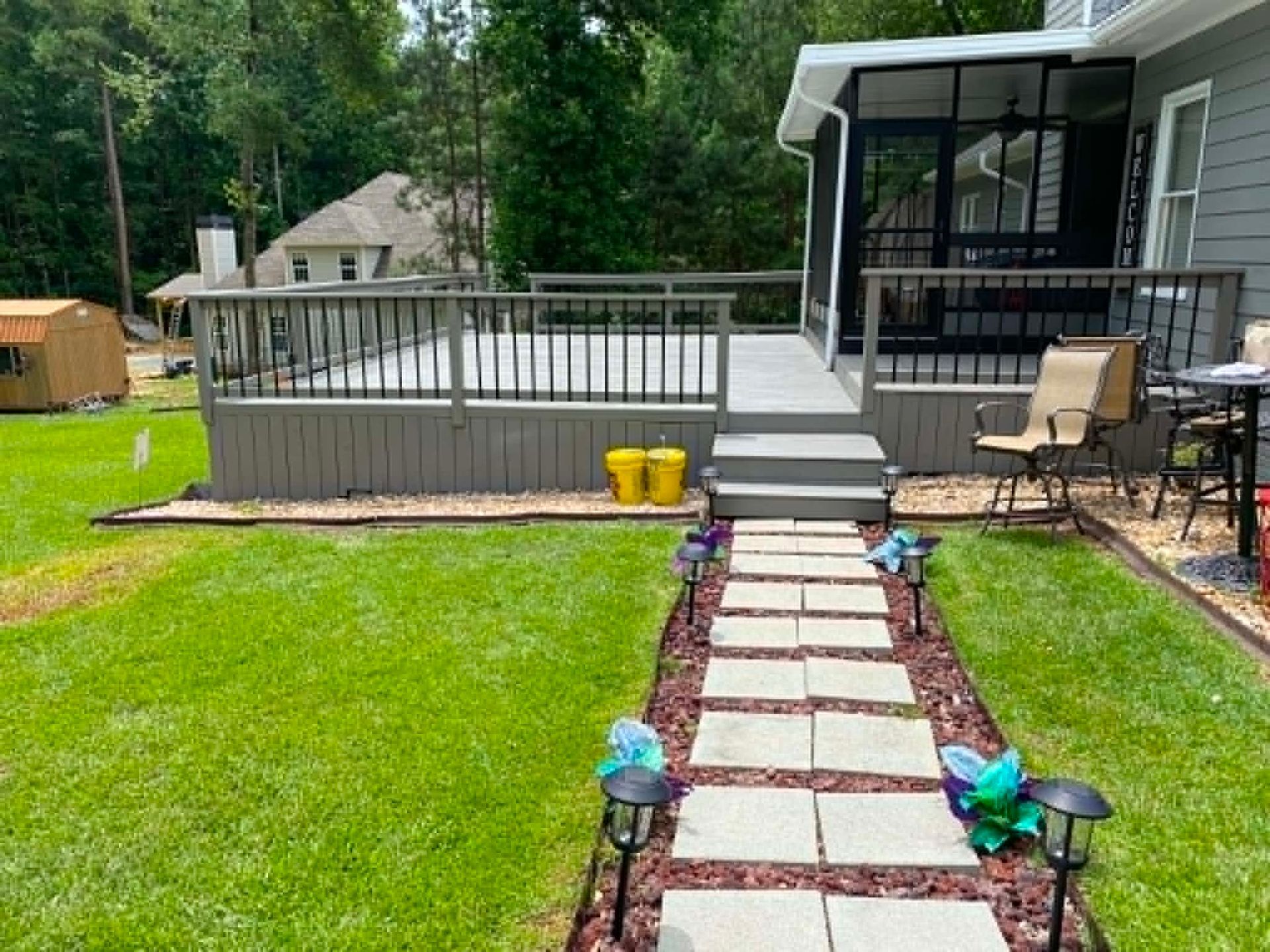 Backyard with stone path, green lawn, gray deck, and patio chairs by the house
