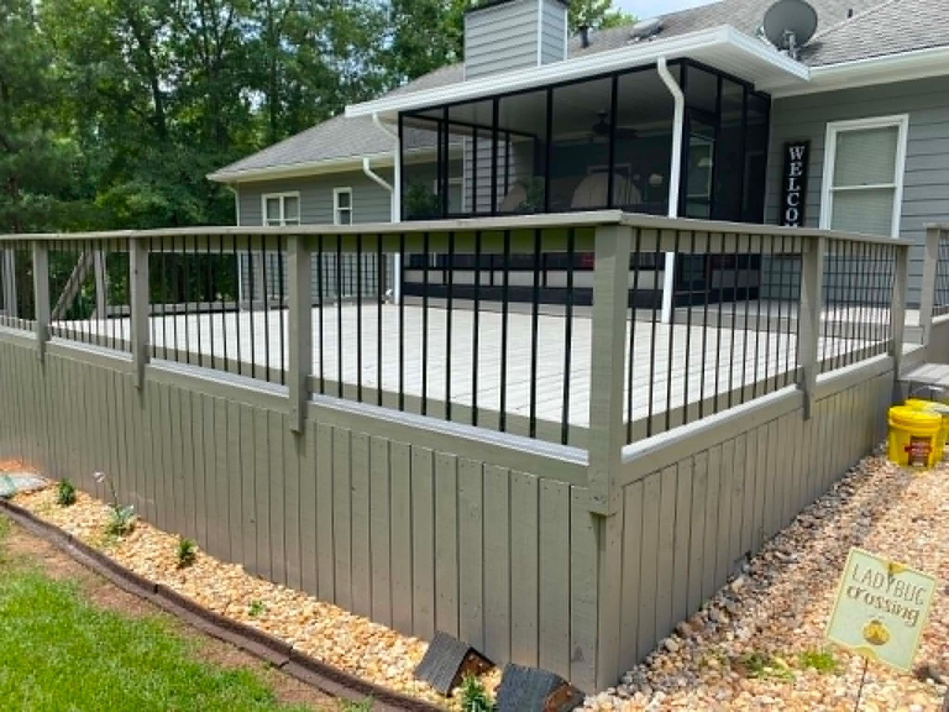 Raised gray deck with black railing beside a house and screened porch, bordered by stone landscaping.