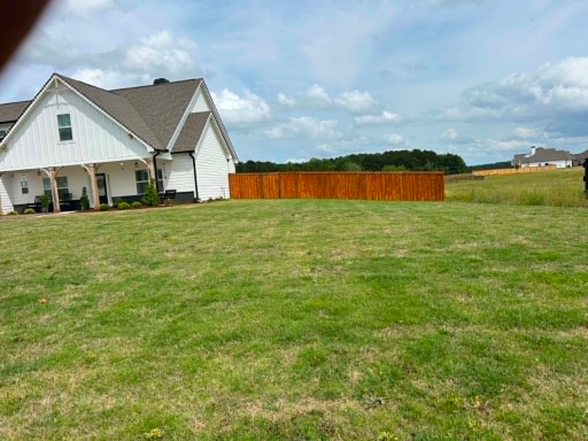 Suburban house beside a large grassy yard with a long orange fence under a partly cloudy sky