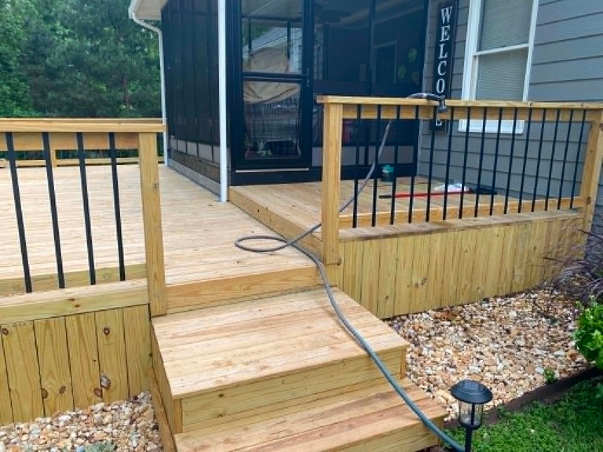 Wooden porch steps with black railing leading to a front deck and screened entry, with a hose on the floor.