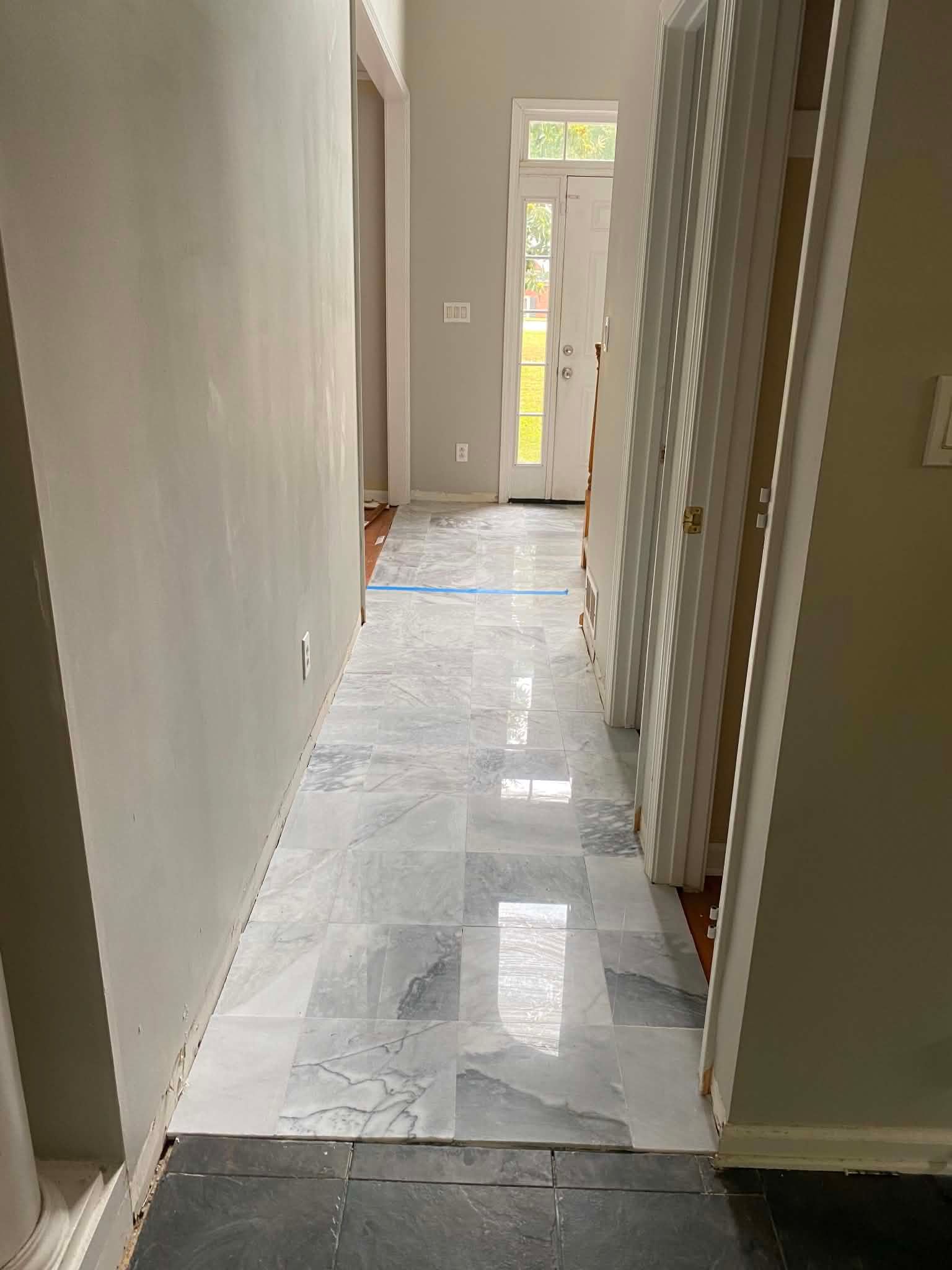 Narrow hallway with marble floor, white walls, and sunlight from a glass door at the far end