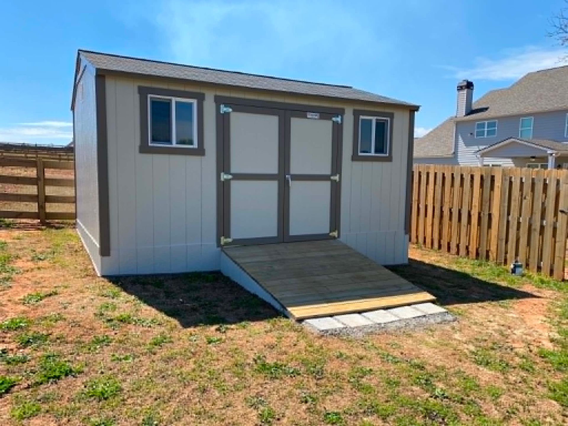 Small gray backyard shed with double doors, ramp, and two windows in a fenced yard