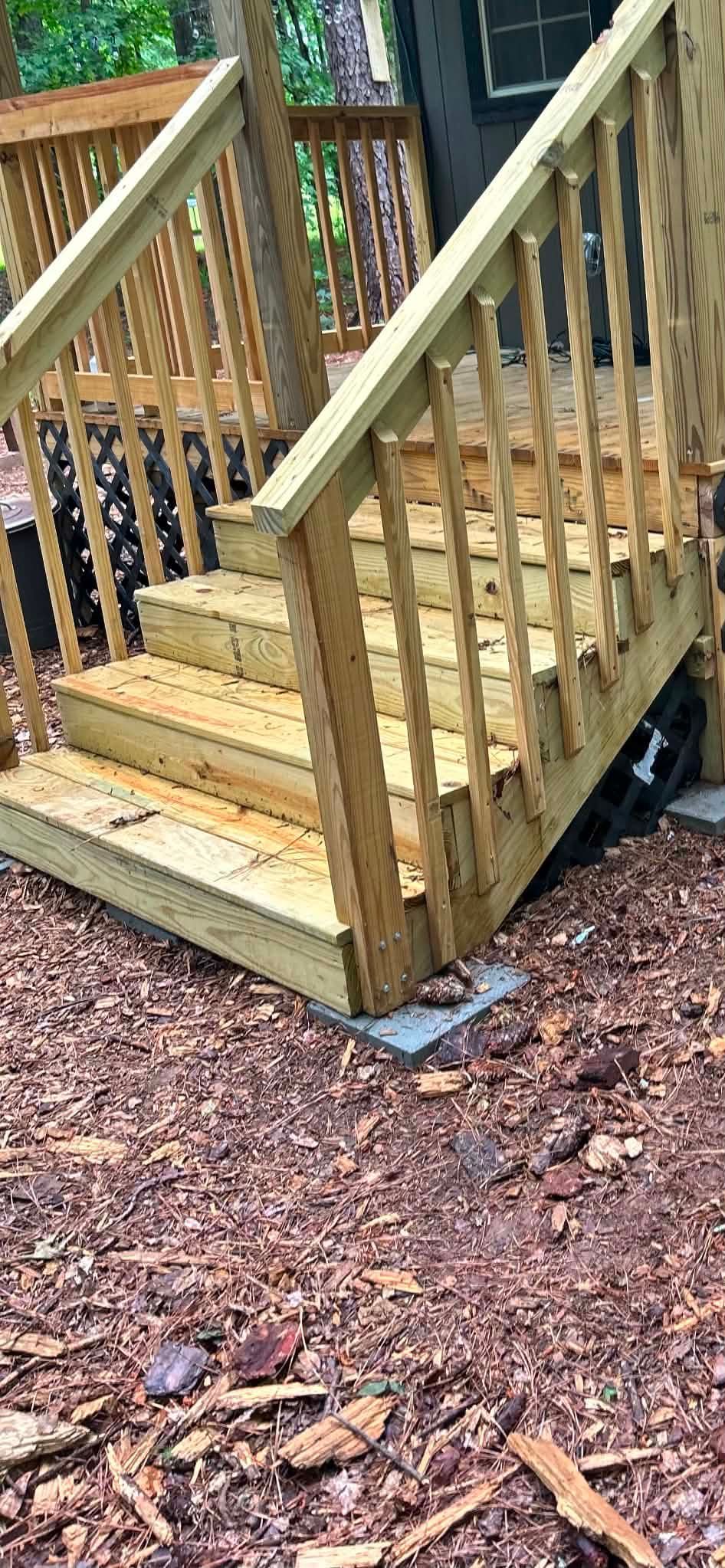 Wooden porch steps with railings leading to a small deck over mulch and leaves.