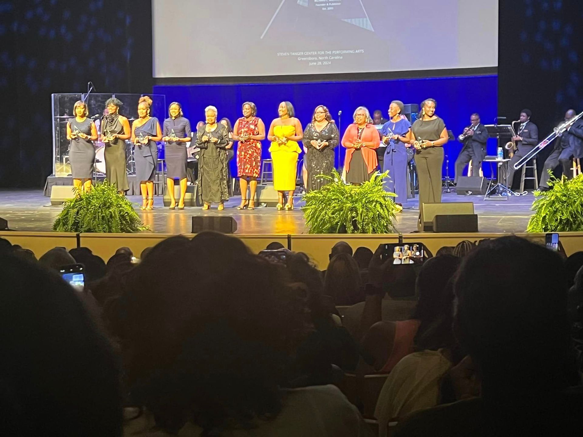 A group of elegantly dressed women on a stage, possibly at an awards ceremony, facing an audience.