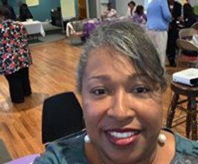 Smiling Black woman in room, wearing earrings, looking at camera. Other people and tables in background.