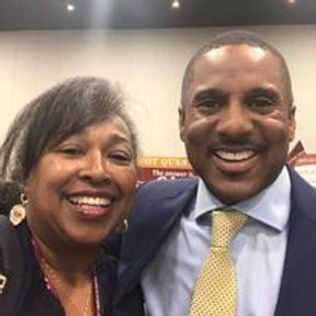 Woman and man smiling, posing for photo. Woman has gray hair, man wears a suit and tie.
