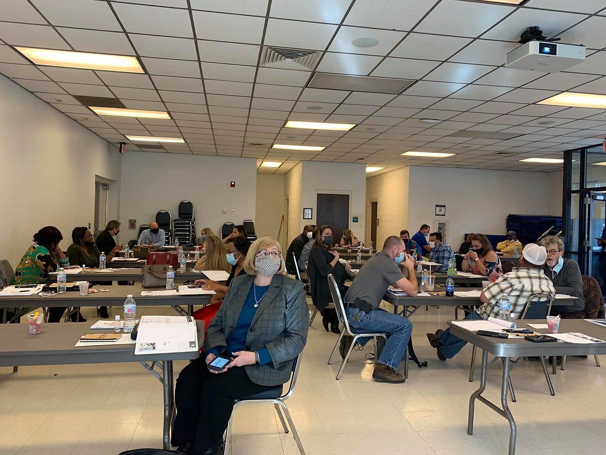 People seated at tables in a large room, some wearing masks. Conference or workshop setting.