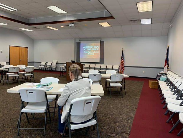 Woman seated at a table in a mostly empty room, facing a projector screen. Tables and chairs are set up for an event.