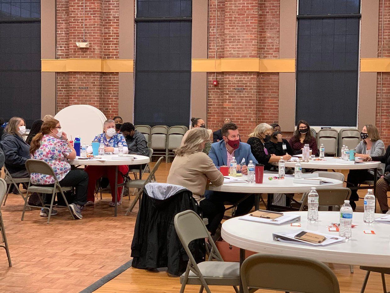 People seated at round tables, masked, in a large room; holding discussions and writing, chairs and tables.