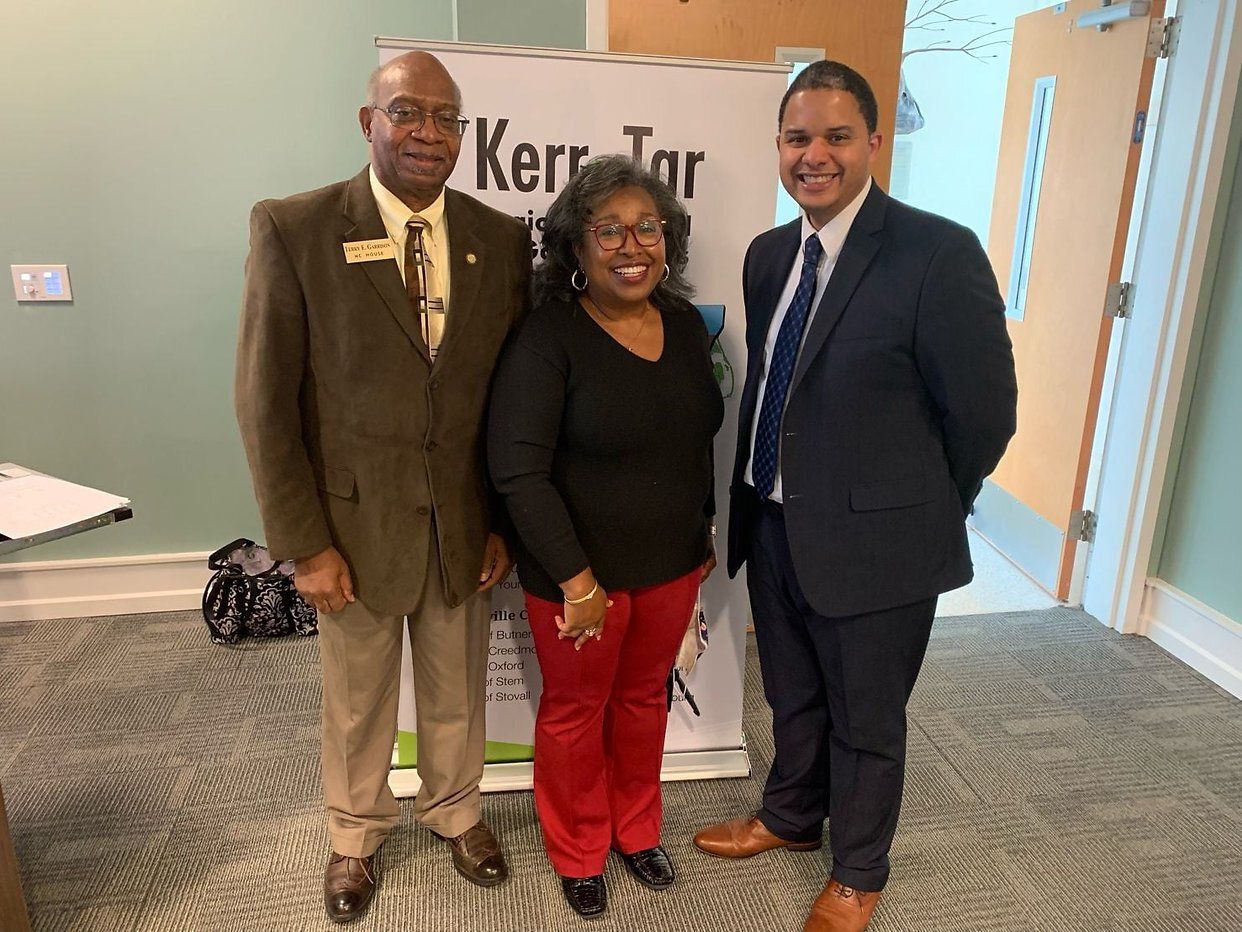 Three people stand by a banner. Man in a suit, woman in red pants, and man in a brown blazer smile.