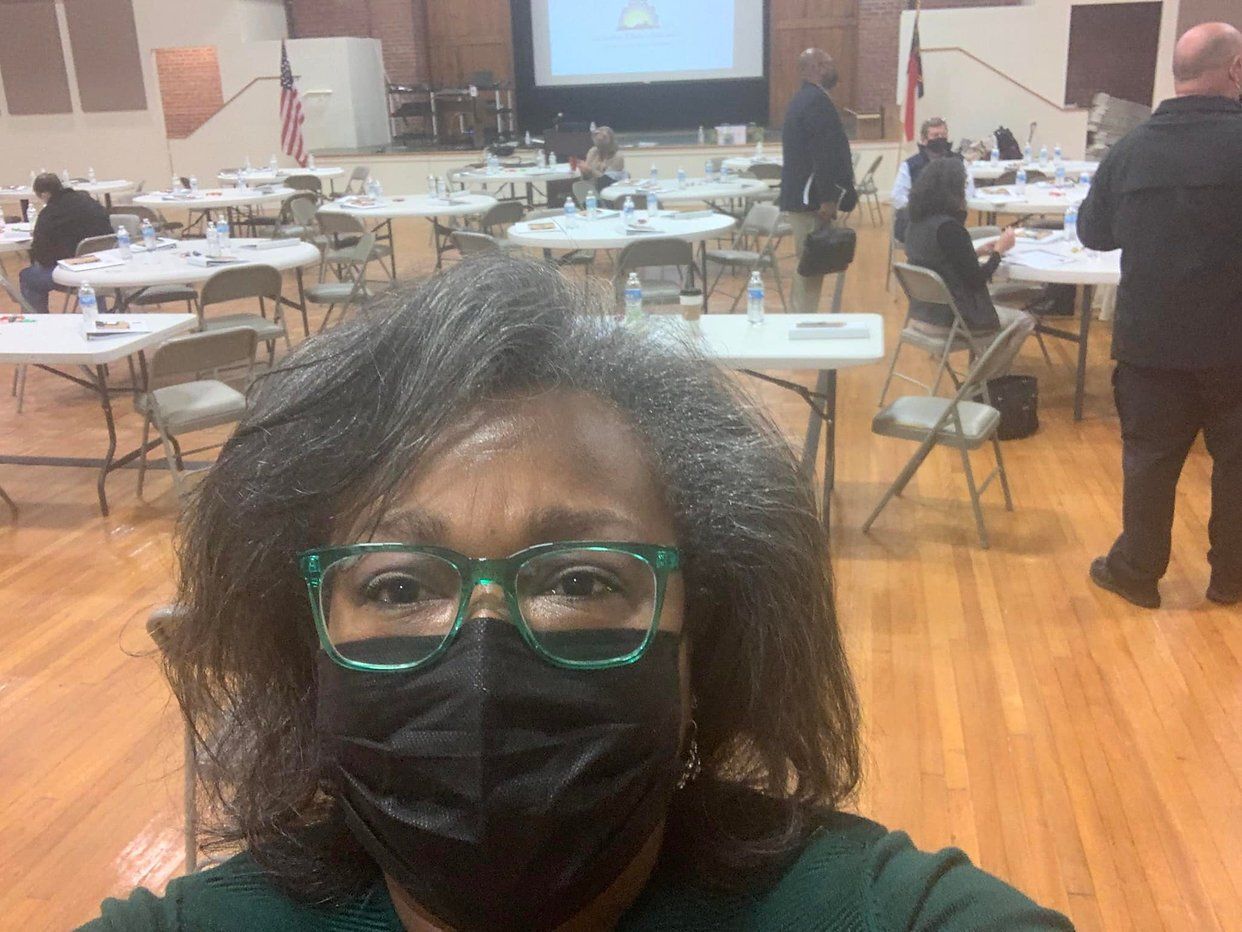Woman in glasses and mask selfies in a room with tables, chairs, and people; a screen at the back.