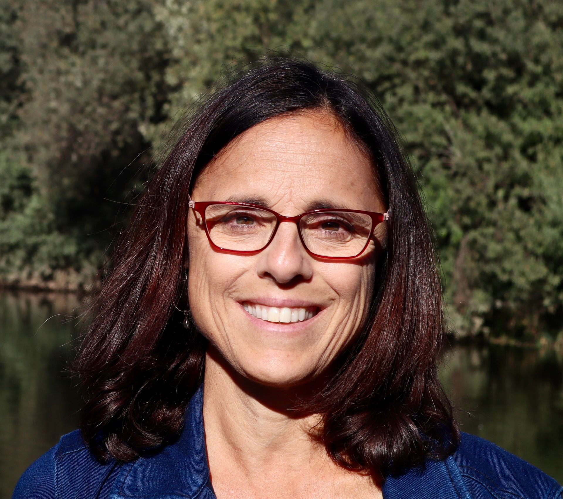 Woman with glasses smiles outdoors, wearing a blue jacket with a park background.