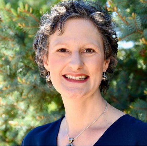 Woman with gray curly hair smiling, wearing a dark blue shirt and silver necklace, outdoors.