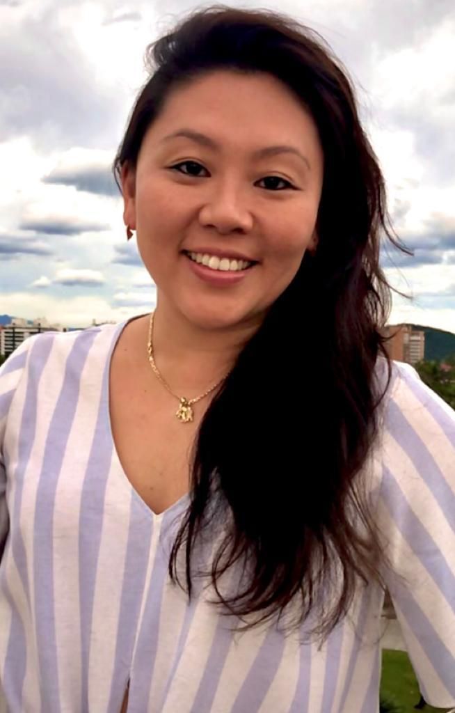 Woman smiles widely outdoors, wearing a striped top and gold necklace. Background shows a cloudy sky and buildings.
