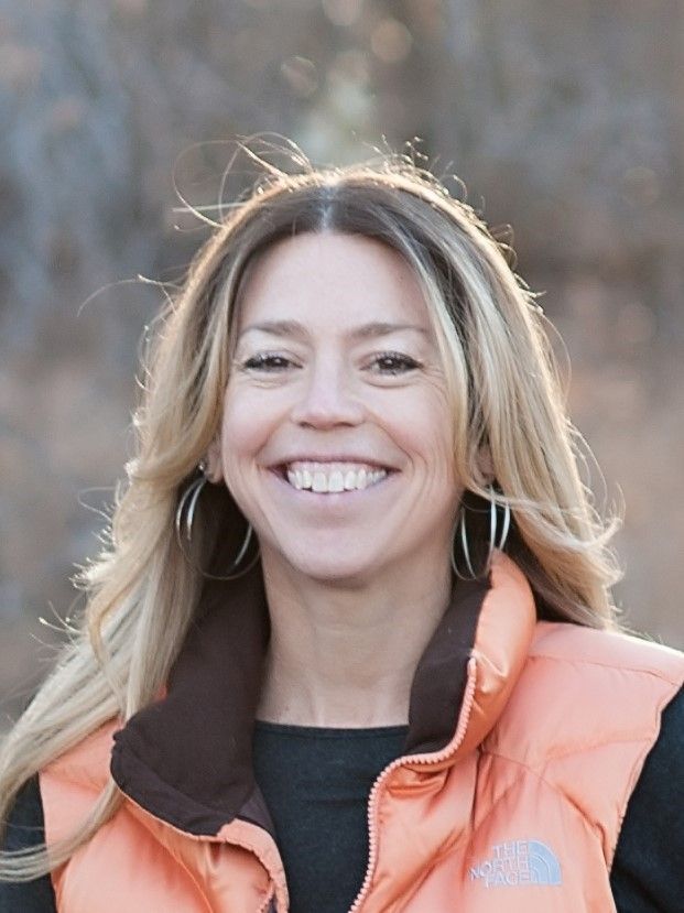 Woman with blonde hair smiles, wearing a light orange vest. Outdoors.