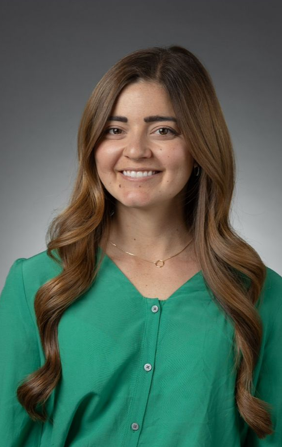 Woman with long brown hair smiles, wearing a green blouse, against a gray background.