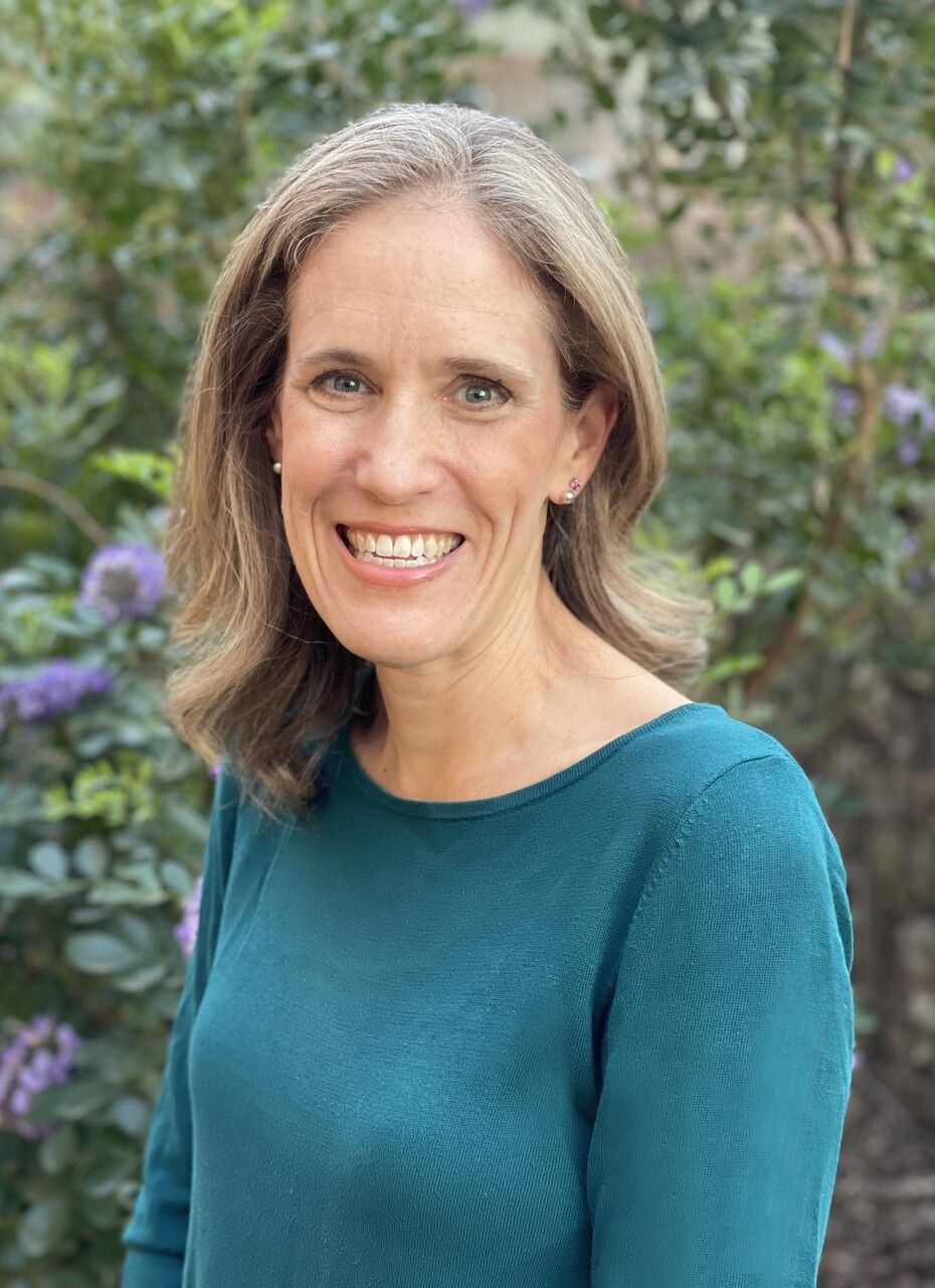 Woman smiling, wearing a teal sweater, standing outside with green and purple foliage in the background.
