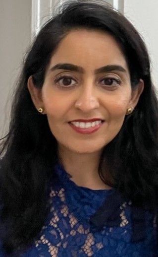 Woman with dark hair smiles, wearing a blue lace top. Indoors, neutral background.