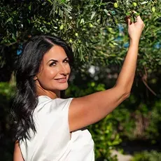 Woman reaching for olives on a tree, smiling; outside in sunny setting.
