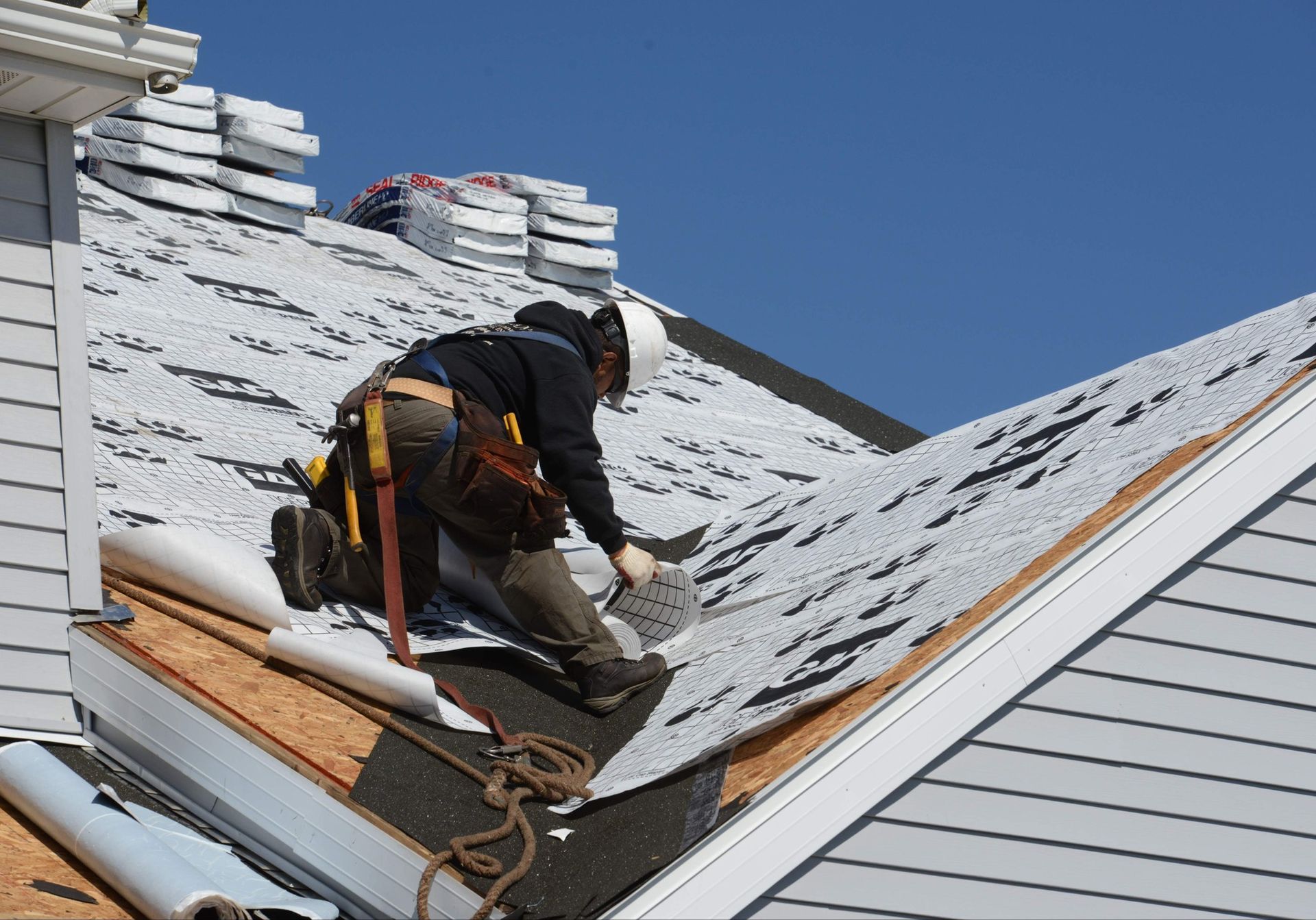 Roofer on a sloped roof, installing shingles. He wears a hard hat and safety gear; blue sky.