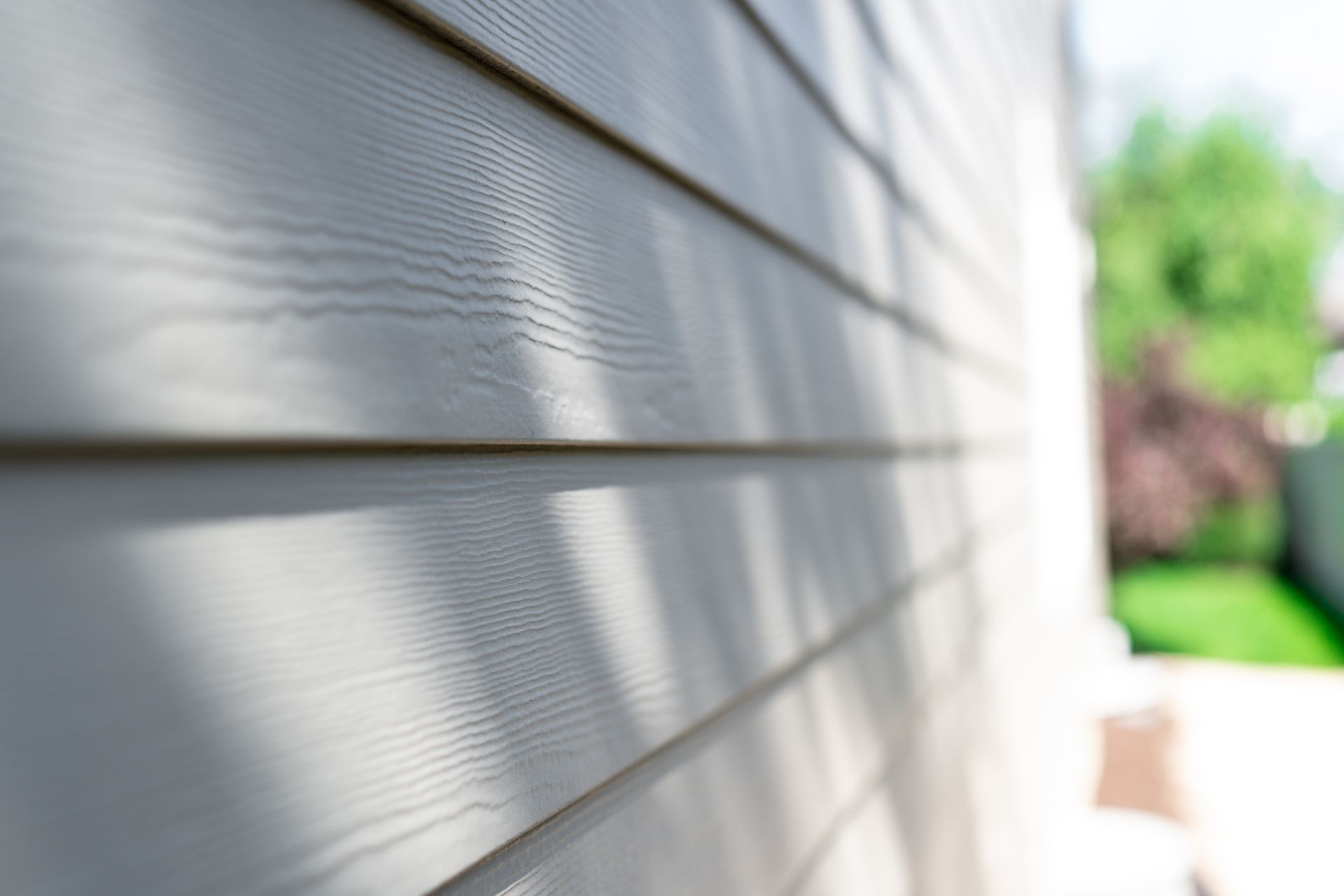 Close-up of light gray house siding, angled with blurred background of greenery and sunlight.