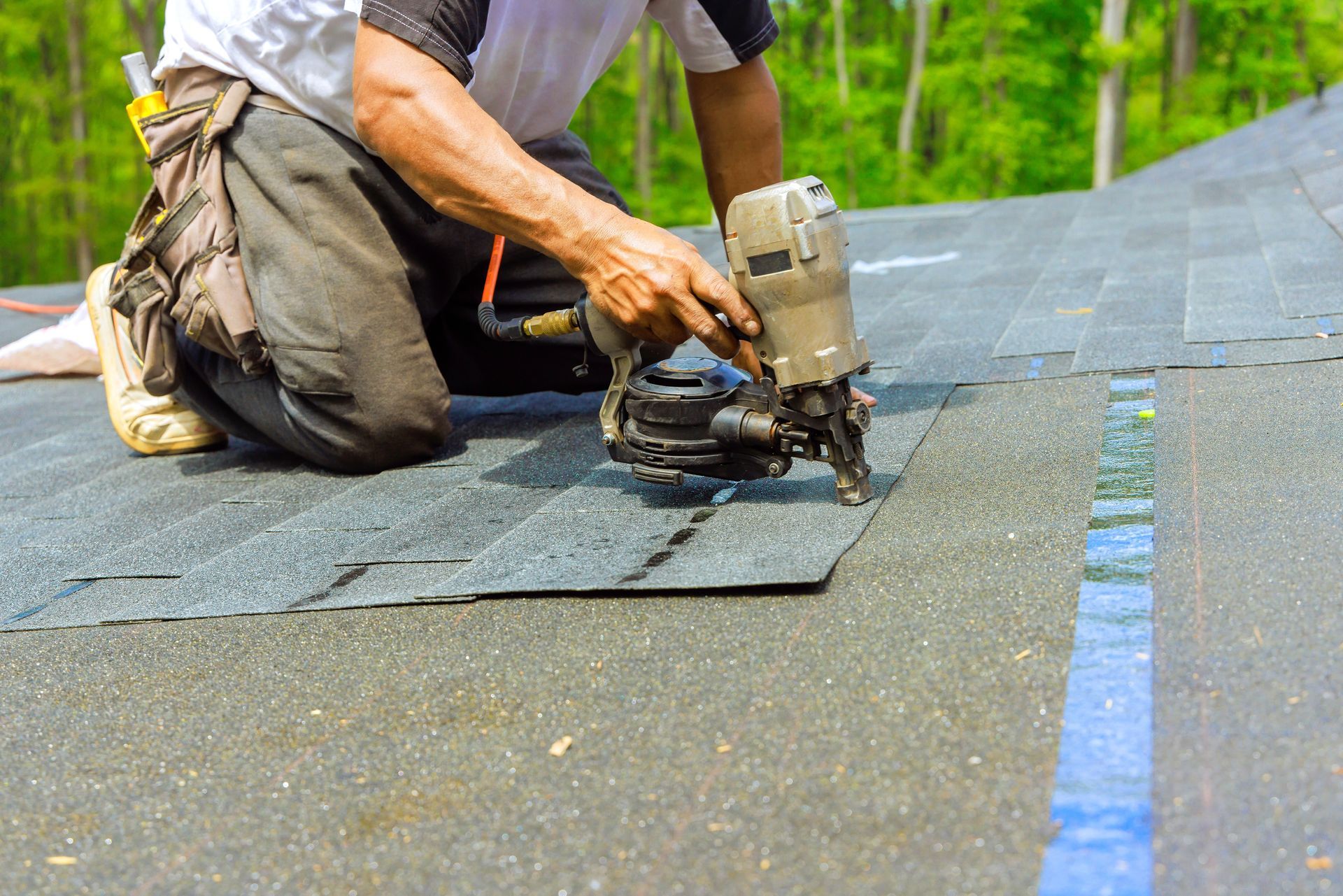 Roofer kneeling, using a nail gun to install asphalt shingles on a roof.