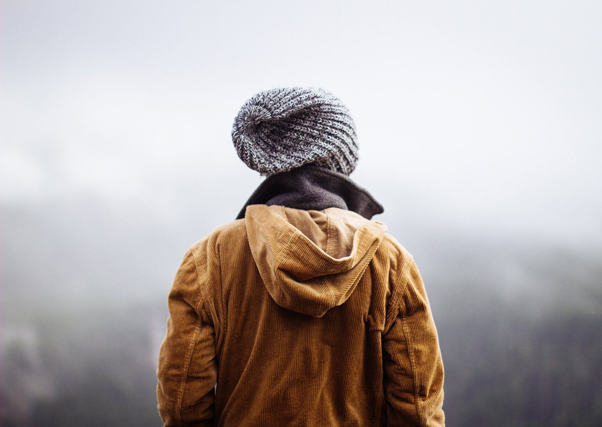 A man wearing a hooded jacket and a knitted hat is standing in front of a mountain.