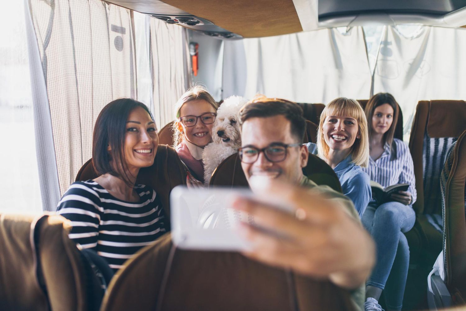 Grupo de pessoas sorridentes e um cachorrinho tiram uma selfie dentro de um ônibus.