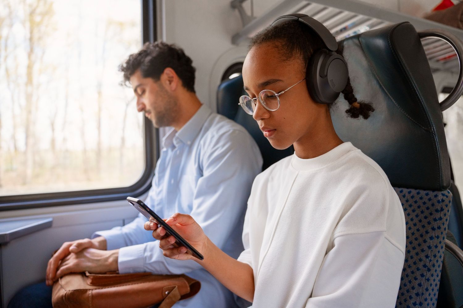 Jovem de óculos e fones de ouvido em um trem, olhando para o celular. Um homem sentado atrás dela.
