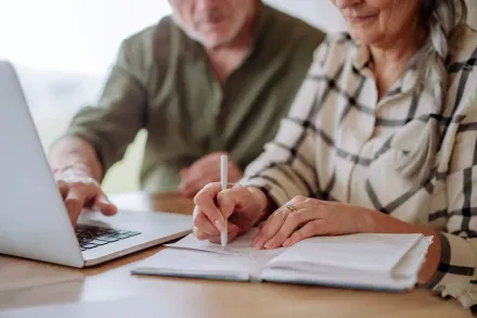 An elderly couple is sitting at a table with a laptop and a notebook.