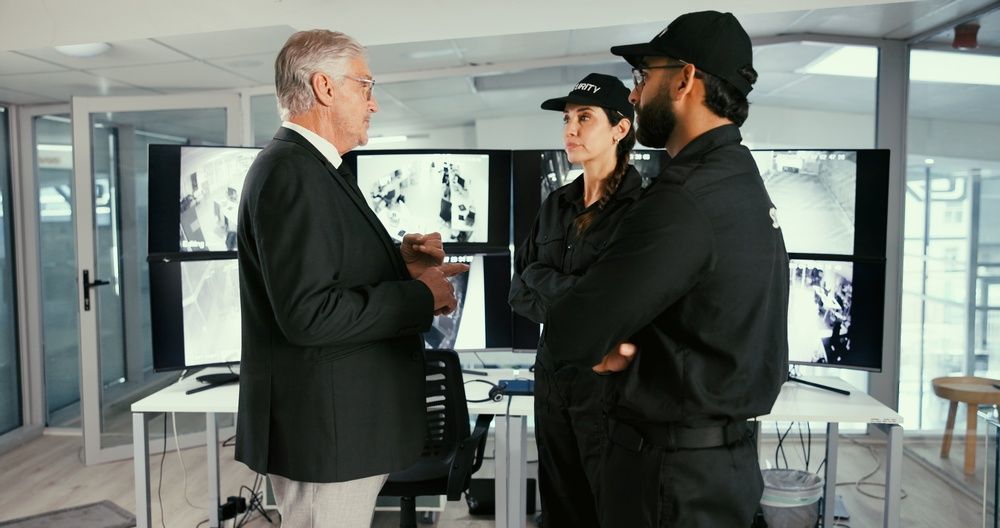 Three people in an office discussing plans, with charts and displays in the background.