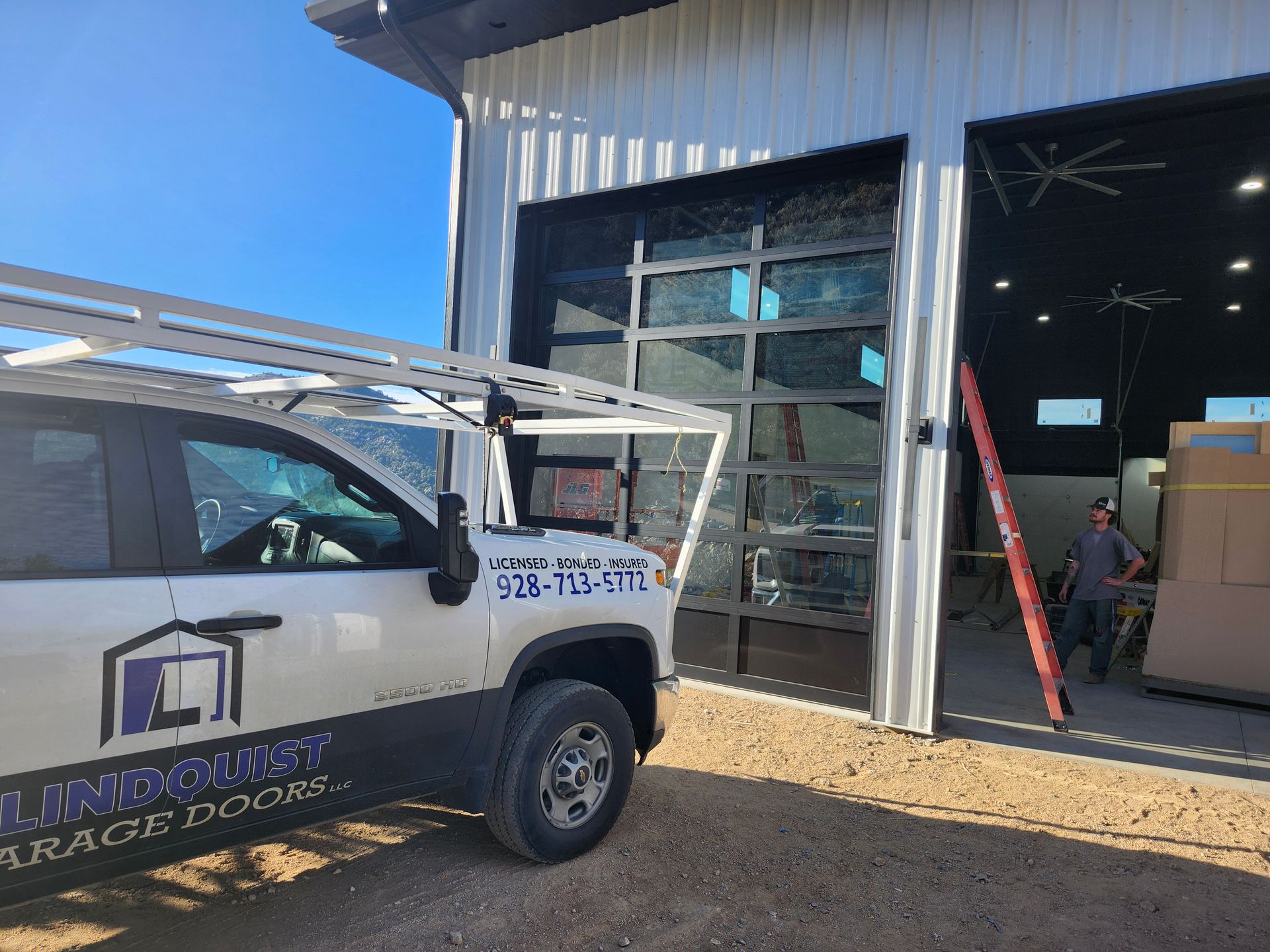 White truck with a ladder rack parked near a building with a large glass garage door. A person stands nearby.