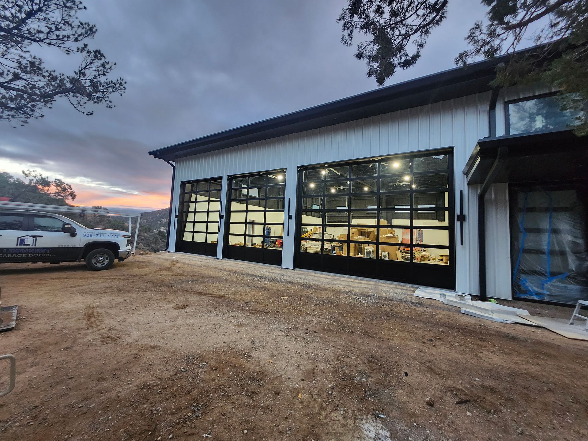Modern white building with black framed glass garage doors, a truck on a dirt lot, sunset.